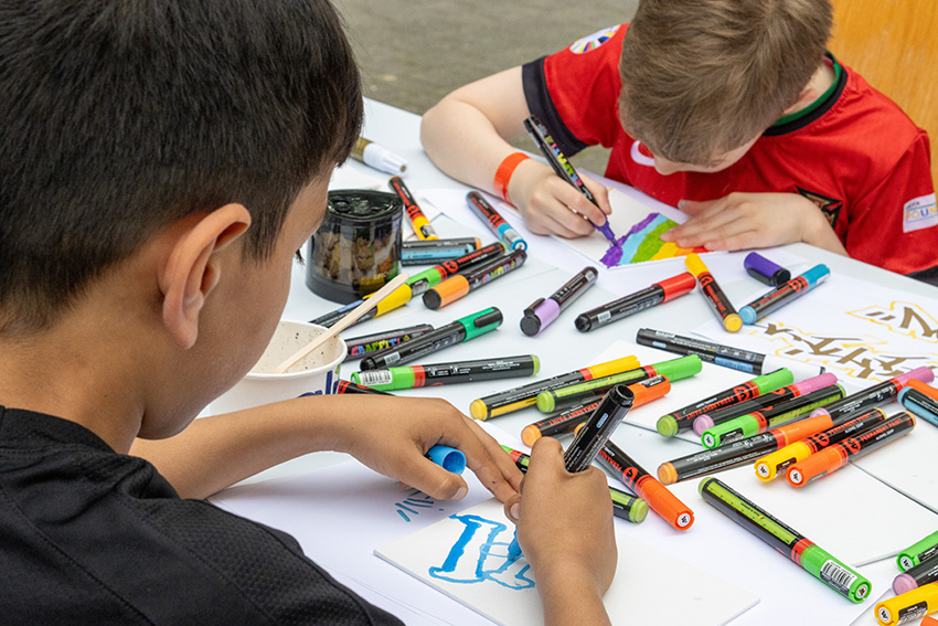 Zwei Kinder sitzen an einem Tisch und zeichnen mit bunten Markern auf weißem Papier. Inmitten der verstreuten Stifte und der konzentrierten Kreativität wird ihr Kunstwerk zu einem automatisch gespeicherten Entwurf der kindlichen Fantasie.