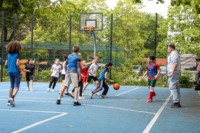 Eine Gruppe von Kindern spielt Basketball auf einem von Bäumen und einem Metallzaun umgebenen Platz im Freien, während ein Erwachsener vom Spielfeldrand aus das muntere Treiben beobachtet - ein automatisch gespeicherter Entwurf von fröhlicher Teamarbeit in Bewegung.