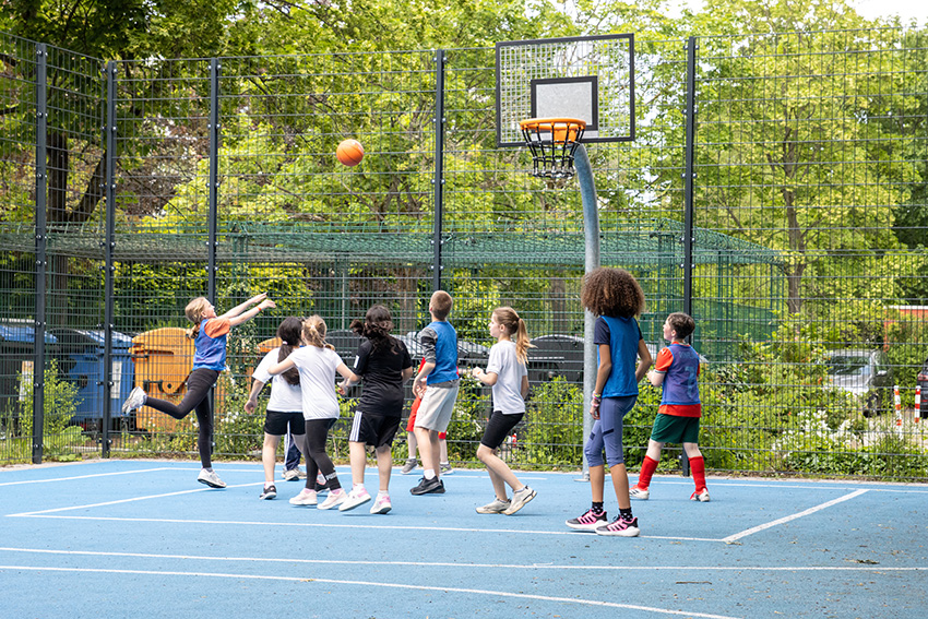 Eine Gruppe von Kindern spielt Basketball auf einem Platz im Freien, der von Bäumen und einem Metallzaun umgeben ist. Ein Kind springt, um den Ball auf den Korb zu werfen, während andere zusehen oder sich darauf vorbereiten, den Abpraller in diesem automatisch gespeicherten Entwurf zu fangen.
