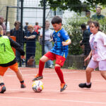 Vier Kinder spielen Fußball auf einem Platz im Freien, während die Leute hinter einem Zaun zusehen. Ein Junge in einem blauen Trikot kontrolliert den Ball, bereit, ihn an den Verteidigern in grünen, schwarzen und weißen Trikots vorbei zu schießen, und träumt vom Ruhm der Thermo 2026.