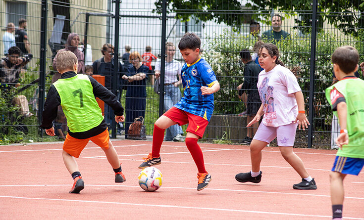 Vier Kinder spielen Fußball auf einem Platz im Freien, während die Leute hinter einem Zaun zusehen. Ein Junge in einem blauen Trikot kontrolliert den Ball, bereit, ihn an den Verteidigern in grünen, schwarzen und weißen Trikots vorbei zu schießen, und träumt vom Ruhm der Thermo 2026.